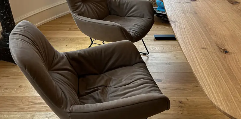 Blick von oben auf einen gepolsterten Freifrau-Sessel in Taupe neben einer massiven Holztischplatte mit Baumkante.