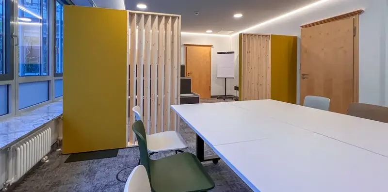 Seminarraum mit langen weißen Tischen, weißen und grünen Schalenstühlen, gelber Akustiktrennwand und Holzlatten-Raumteiler am Fenster.