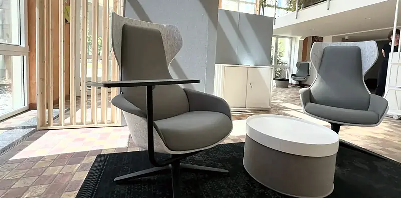 Helle Lounge mit grauem Ohrensessel mit Ablagetisch, weißem Hocker und Holzlatten-Raumteiler am Fenster.