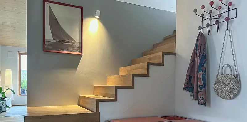 Beleuchtete Eichentreppe an grauer Wand mit Segelbootbild, rechts eine Wandgarderobe mit bunten Kugelhaken.