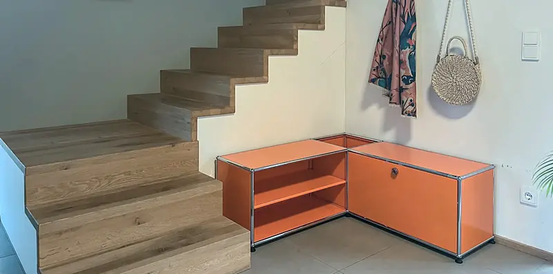 Orangefarbenes USM-Eckmöbel mit offenem Regalfach und Klappe neben den unteren Stufen einer Eichentreppe.