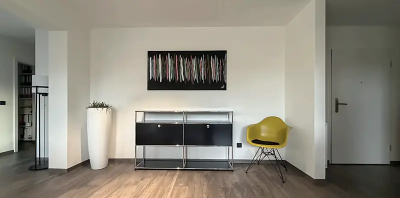Wohnraum mit schwarzem USM-Sideboard mit Chromgestell, gelbem Vitra-Schalenstuhl und weißem Pflanzkübel vor heller Wand.